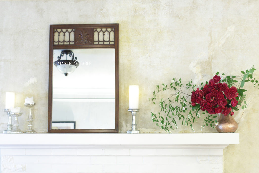 Red roses, mock orange branches in a copper pot on white mantel. Silver candle holders and brown filigree mirror.
