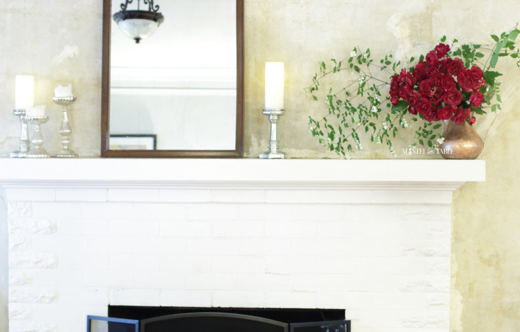 Red roses, mock orange branches in a copper pot on white mantel. Silver candle holders, glowing white candles, and brown filigree mirror.