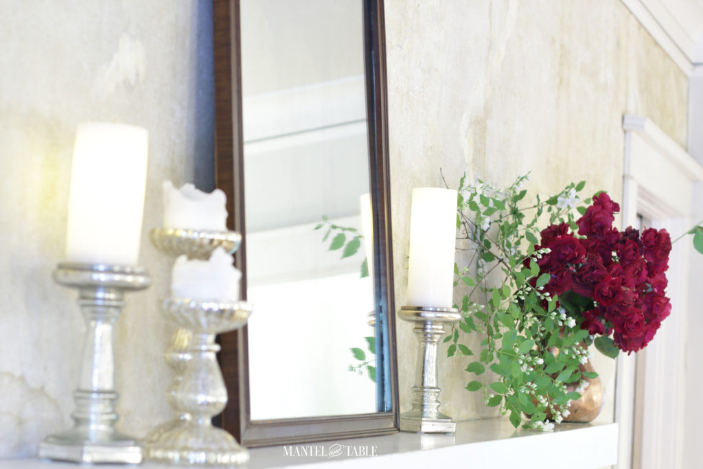 Red roses, mock orange branches in a copper pot on white mantel. Silver candle holders, glowing white candles, and brown filigree mirror. Angle view