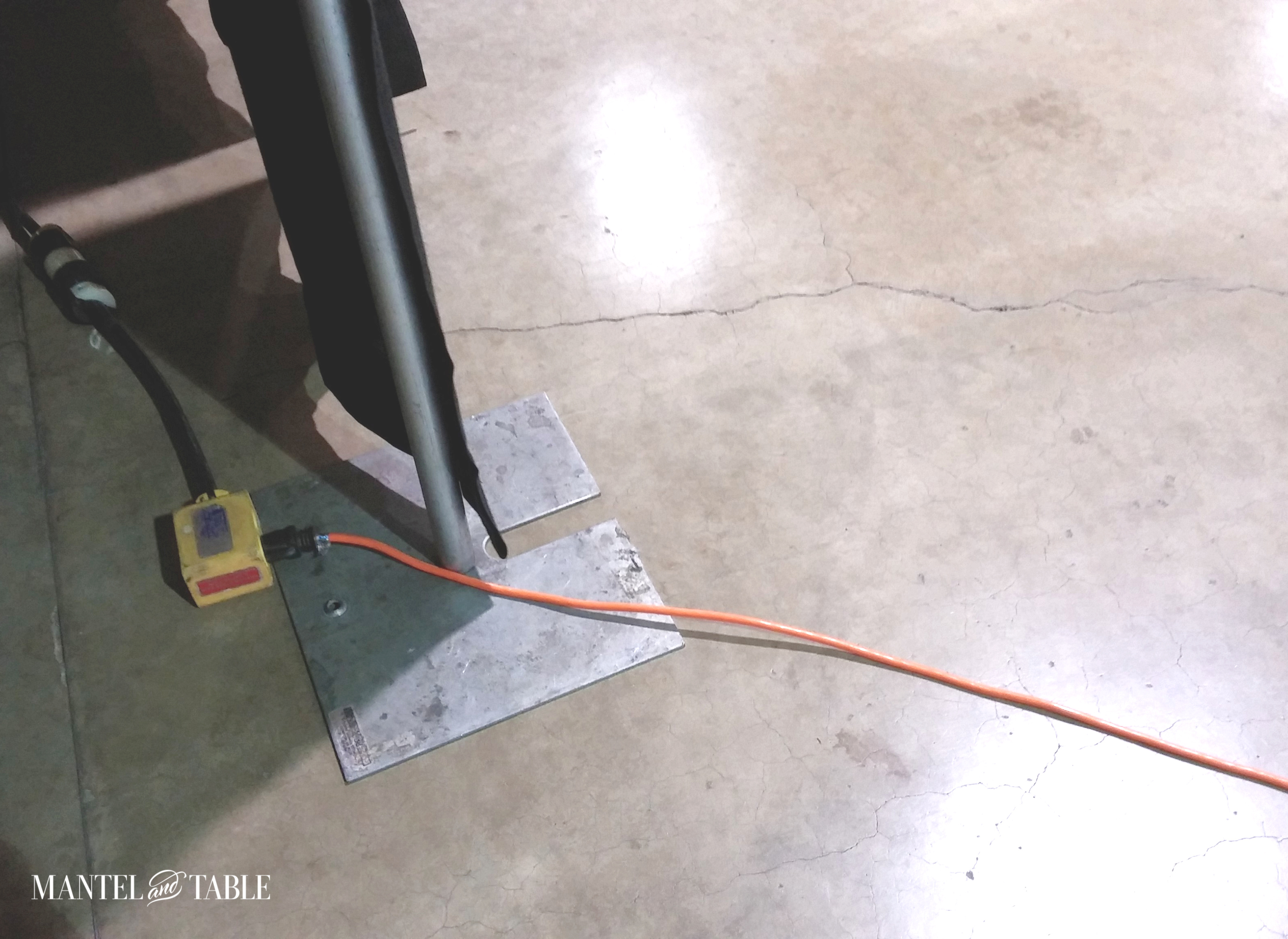 Little electrical outlet box on the floor behind a post. Orange extension cord plugged in