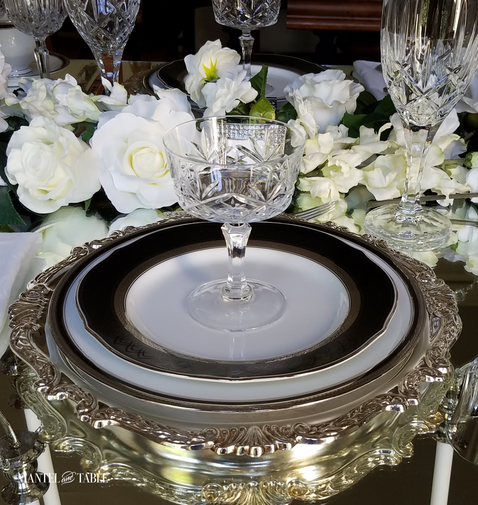 Top 10 Favorites - ornate silver, black, & white place setting on mirrored tabletop. Crystal glasses and white flowers