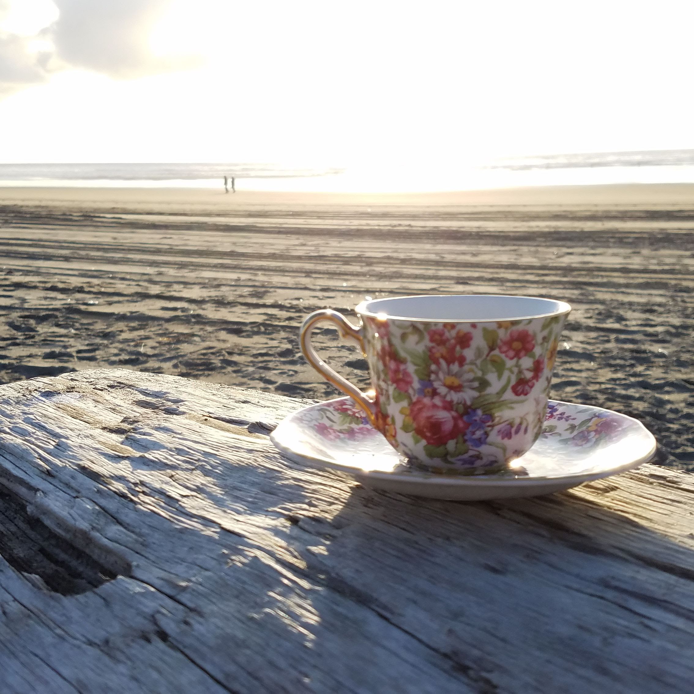 Brightly flowered teacup on driftwood at the beach. Sunset with two people sillouetted in the distance