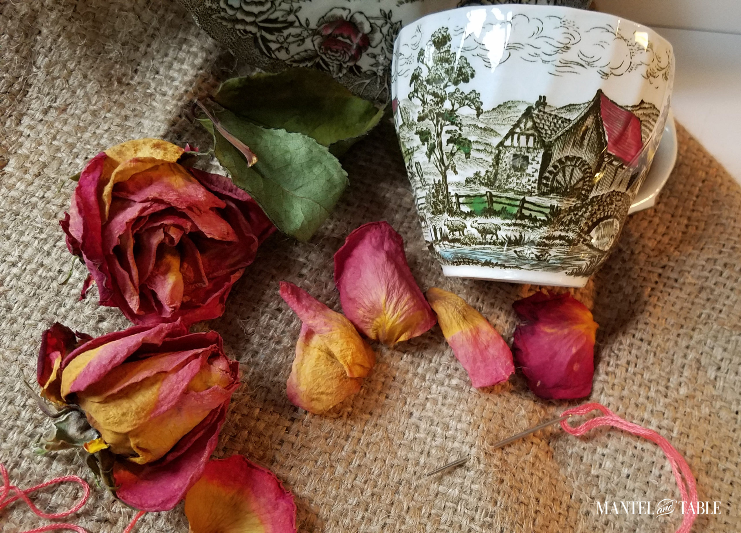 Teacup on its side with burlap, needle & pink embroidery floss, & dried roses