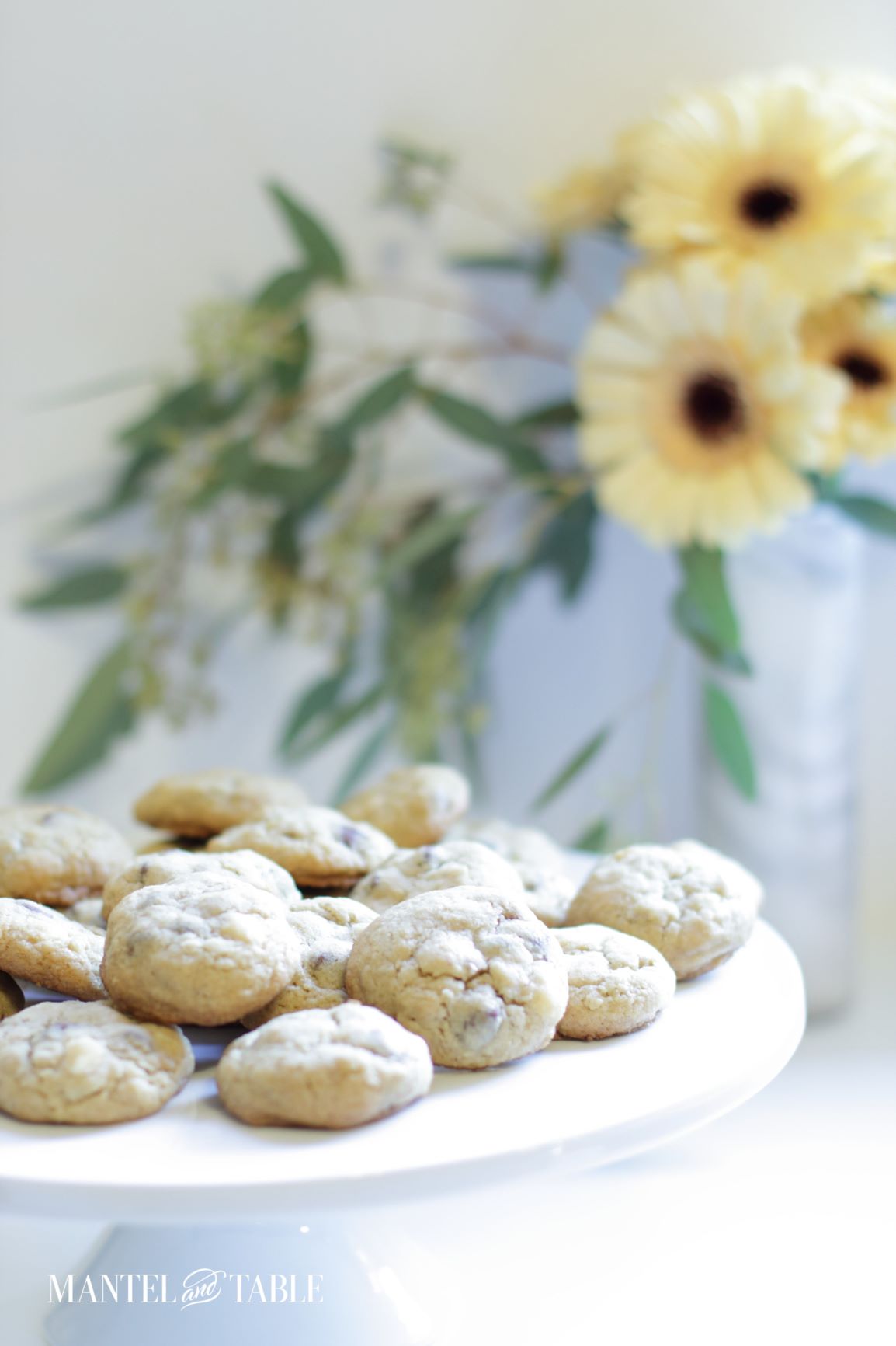 Best Chocolate Chip Cookies ~ baked cookies on a white stand. Pale yellow flowers in the background