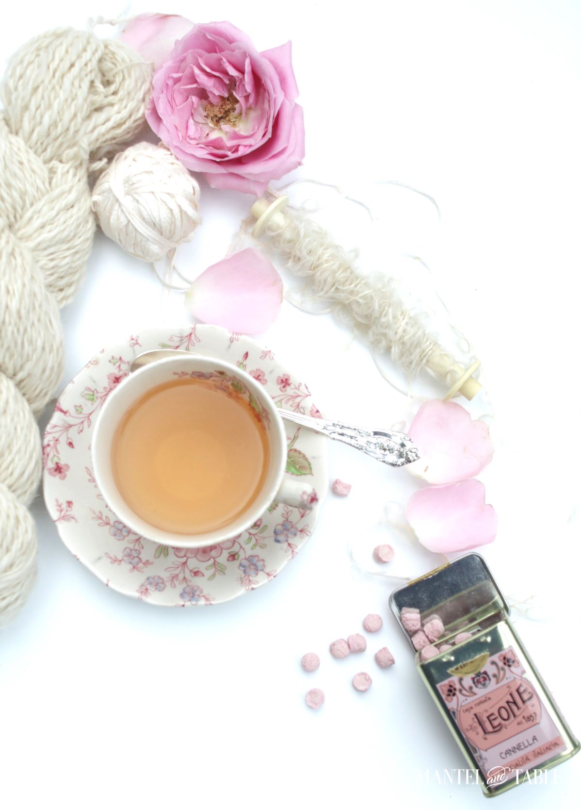 Cropped image of pink flowered teacup flat lay with white yarn, pink rose petals, and tin of pink Leone candies