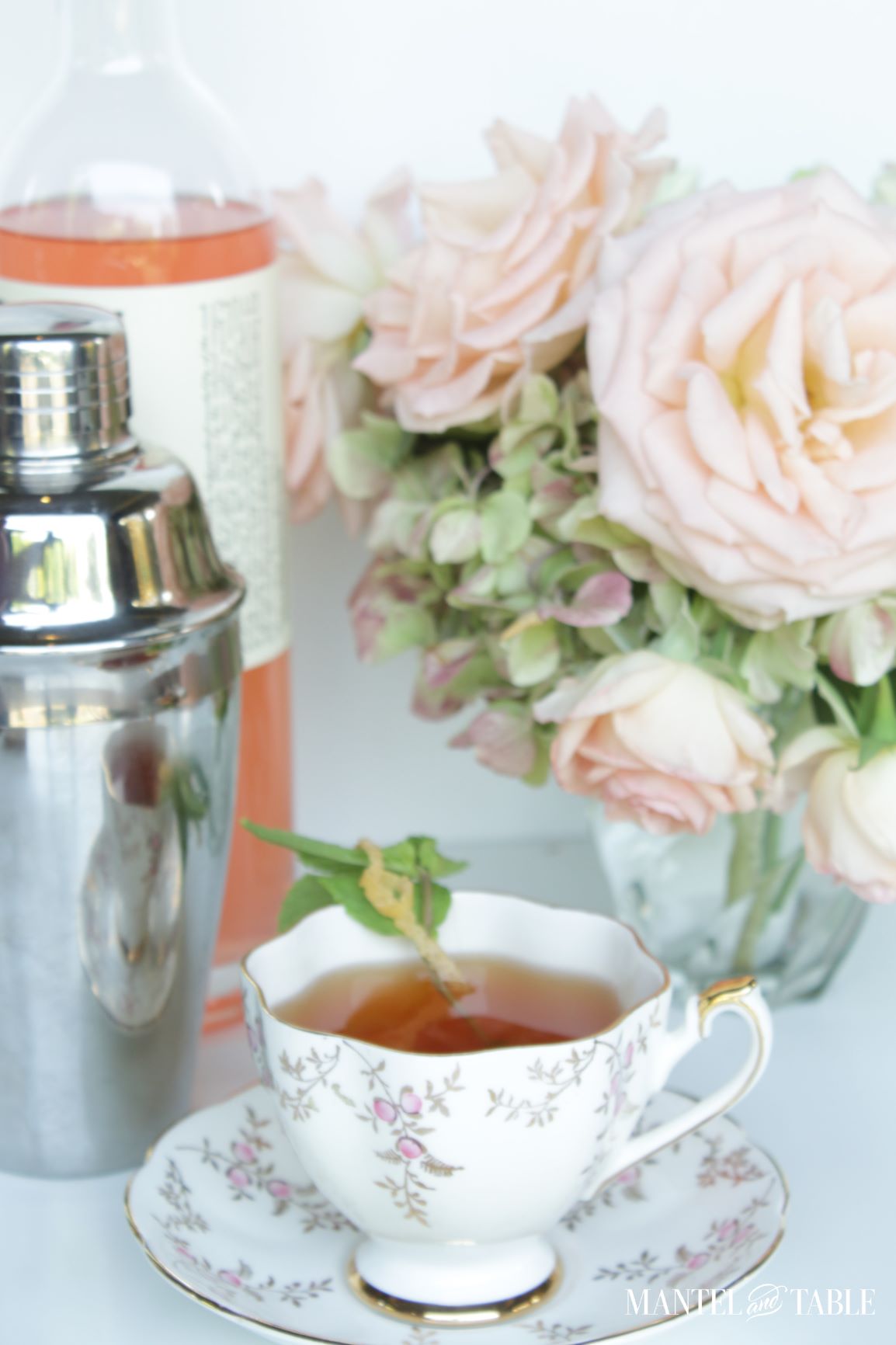 Cocktail in a teacup, silver shaker, grapefruit vodka bottle, pink roses