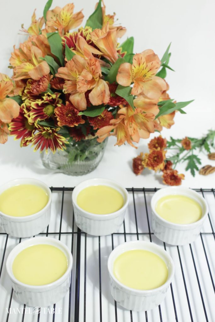 Five ramekins of Pumpkin pot de creme cooling on wire rack. White background and rust/orange flowers