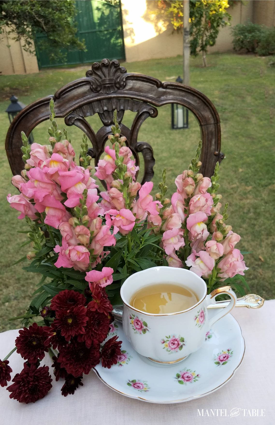 Teacup on edge of table, pink snapdragons on a chair behind