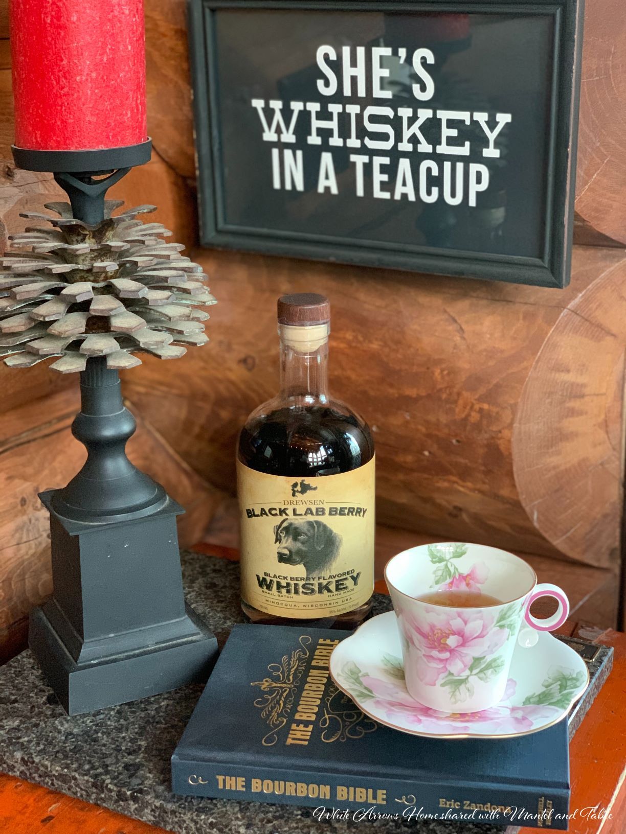 Teacup on table with book, whiskey bottle and lamp under "She's Whiskey in a Teacup" sign