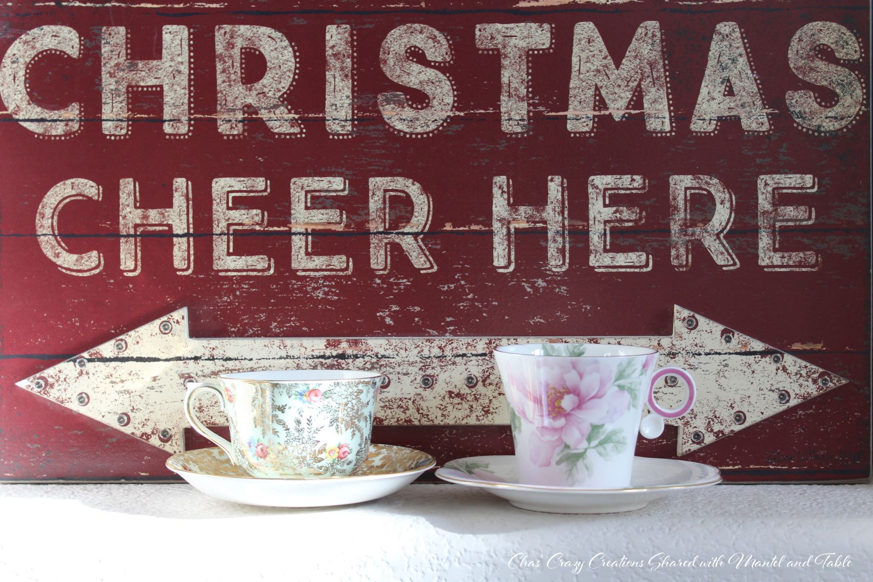 Two teacups and saucers in front of red and white 'Christmas Cheer Here' sign