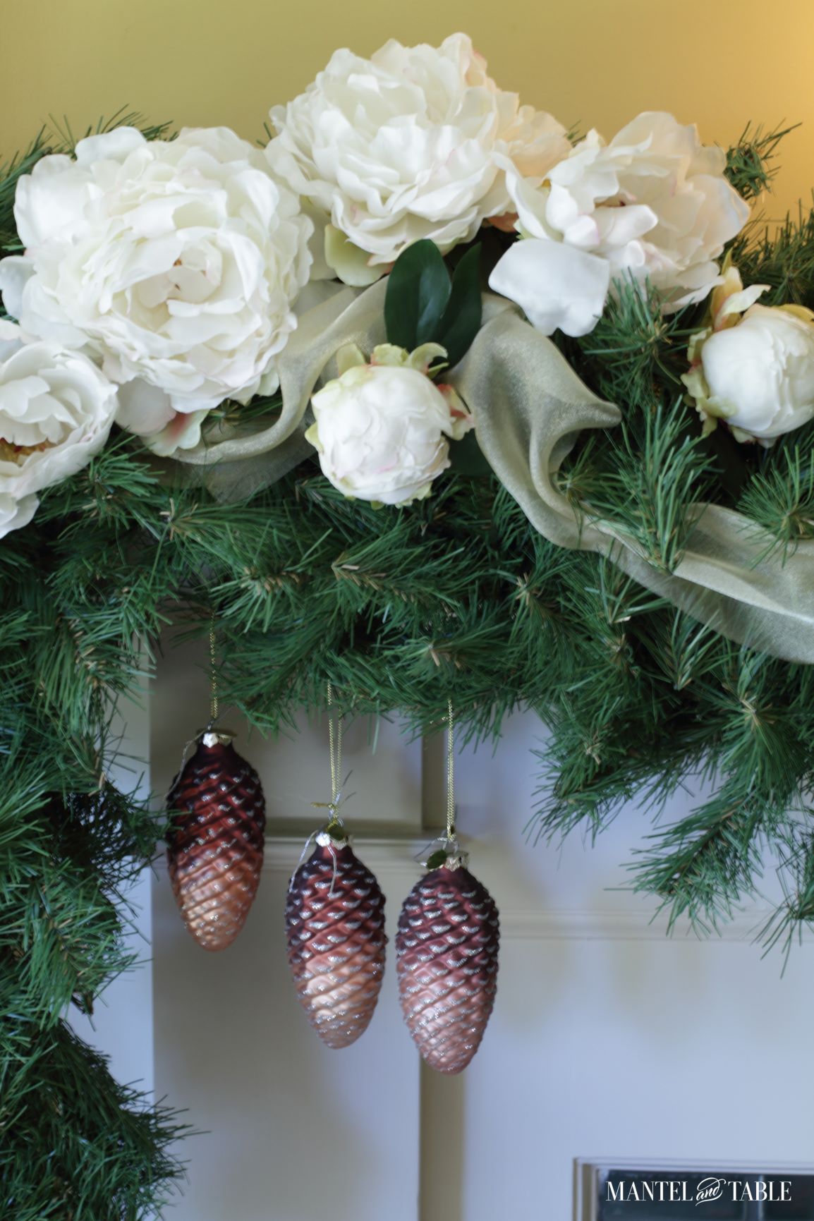 Blush pinecone ornaments hanging from evergreen and white peony mantel garland