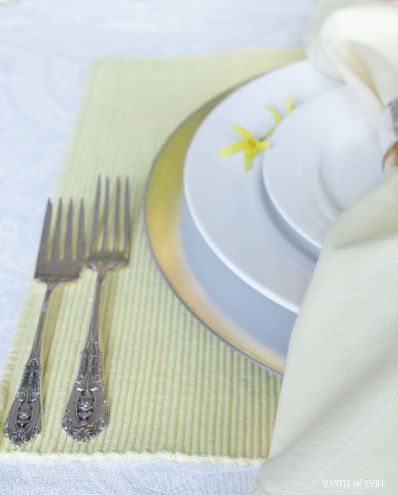 Closeup of silver forks beside white and silver plate stack, yellow napkin in silver ring