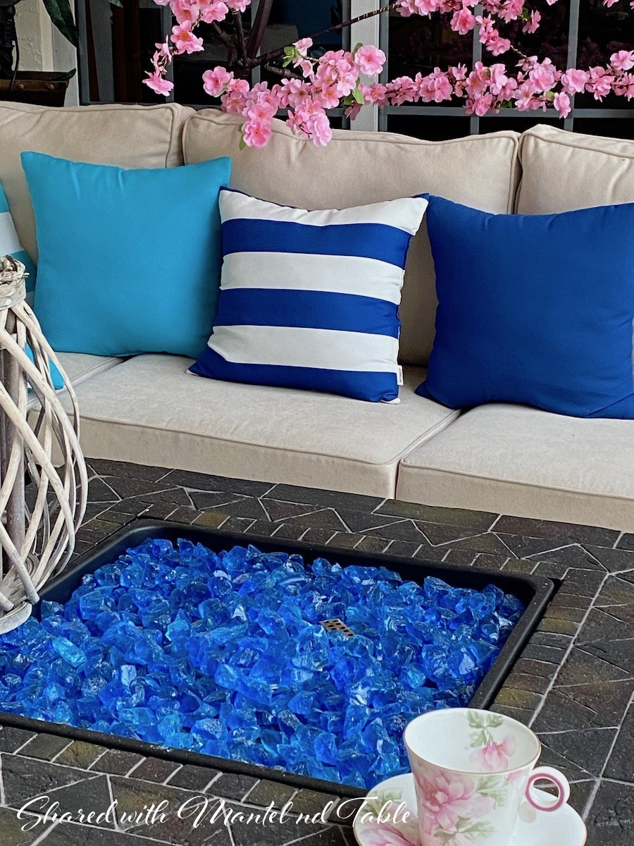 Pink flowering branch overhanging white outdoor couch with blue pillows, blue rock firepit, and charming traveling teacup