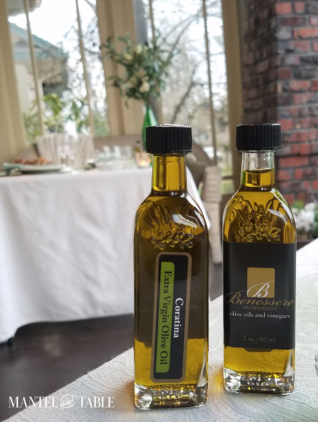Closeup of two small olive oil bottles on white tablecloth, with another similar tablescape in the background