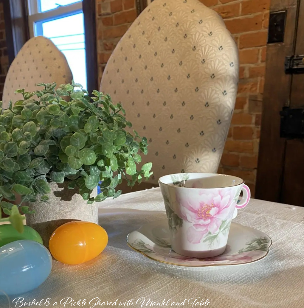 Closeup of teacup and saucer on white tablecloth. Plastic Easter eggs and potted plant