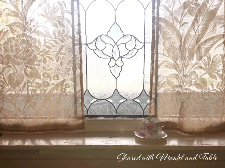 Pink flowered teacup and saucer on cream colored window still, leaded glass window and floral lace curtain panels
