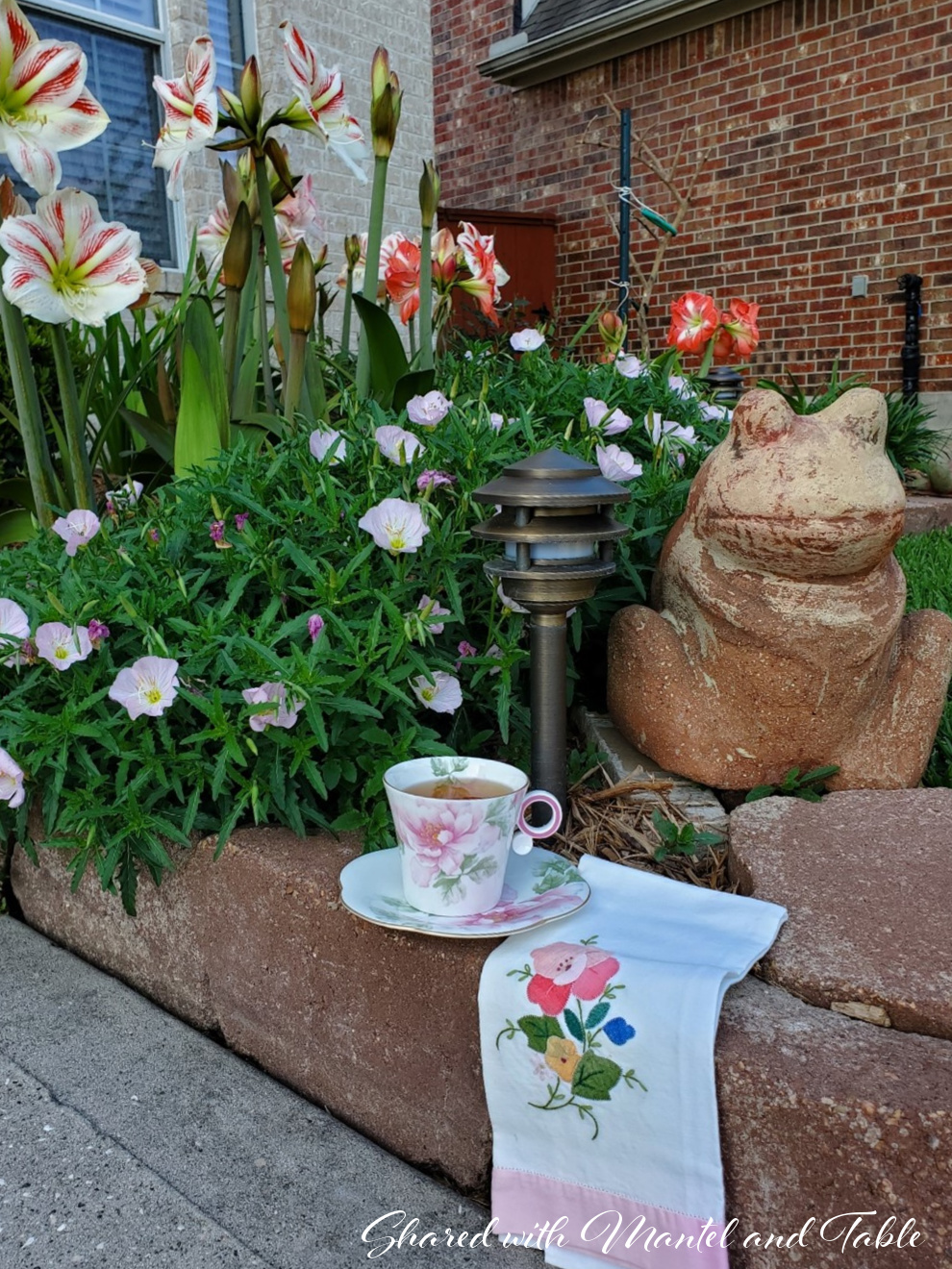 Pink flowered teacup and saucer on the brick border of flower garden. Embroidered tea towel and concrete garden frog statue