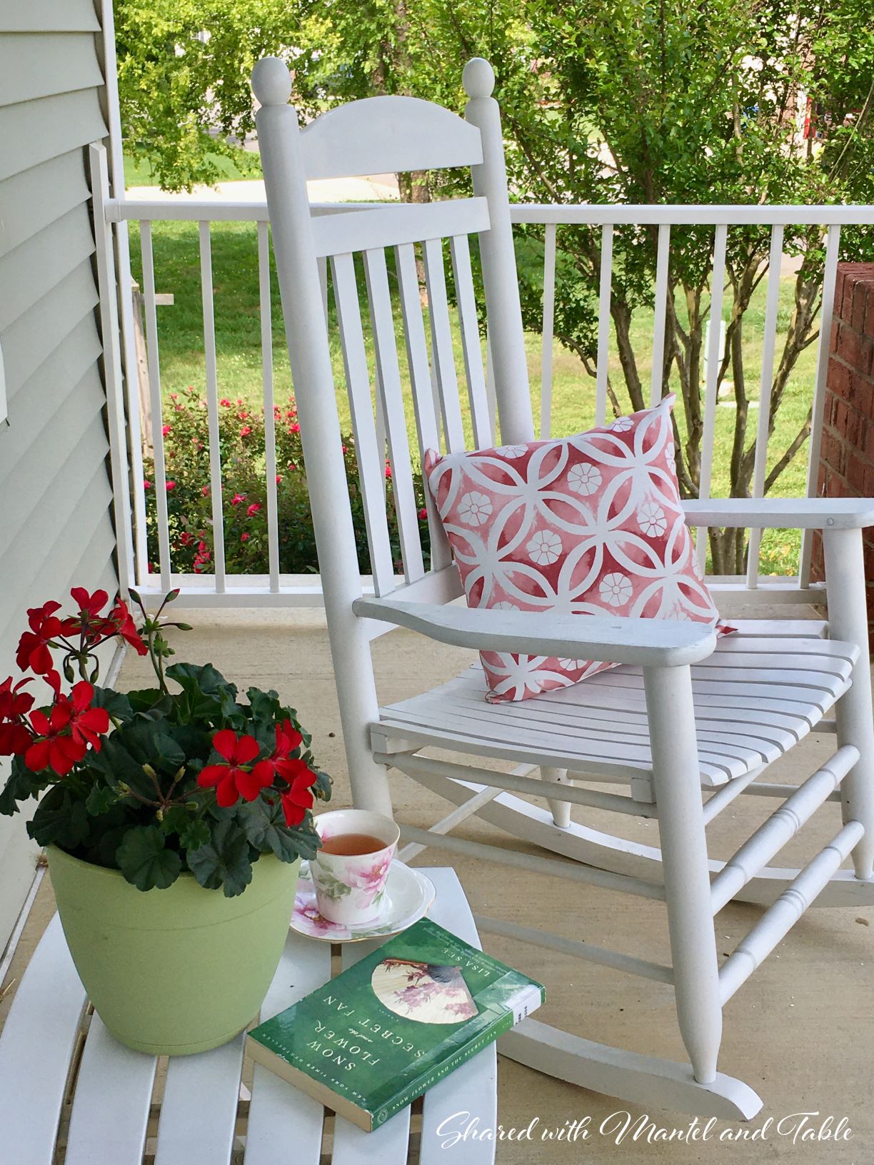 White rocker on porch with teacup on table with book and red geranium