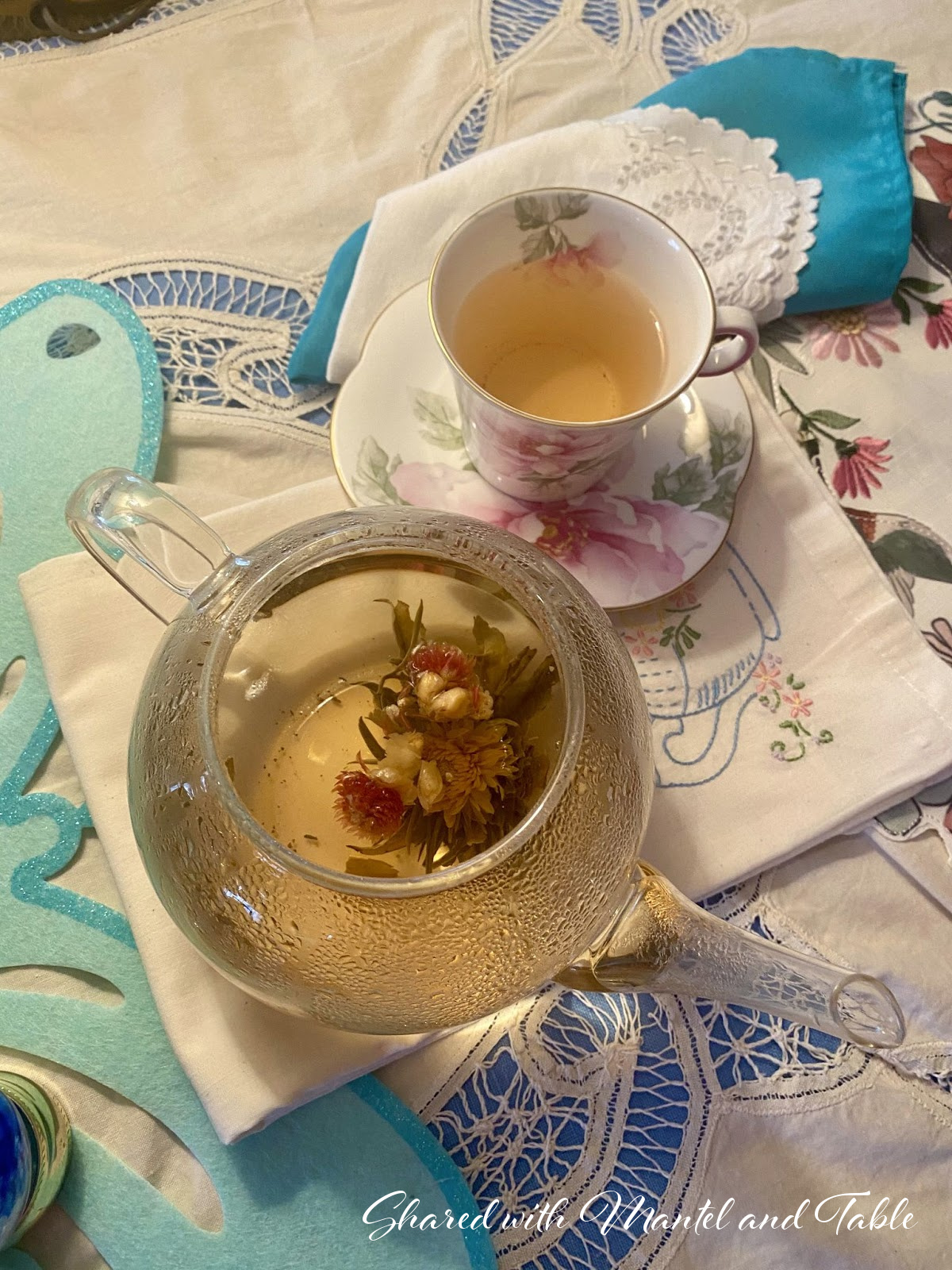 Pink & white flowered traveling teacup next to a clear teapot with flowers in the tea