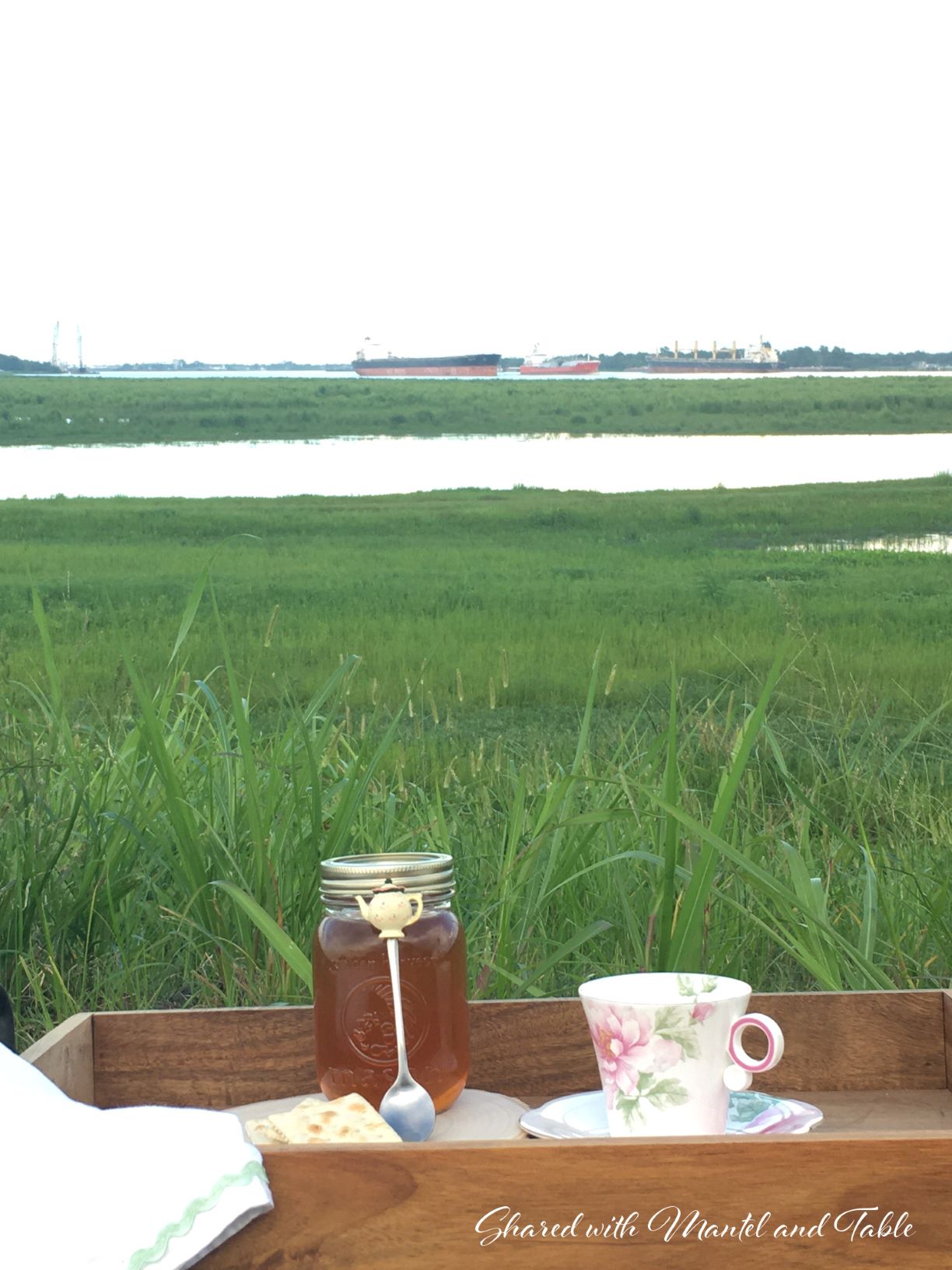 Wooden tray holding teacup, jar of honey sitting in the grass beside the Montz Louisiana spillway. Three ships in the distance.