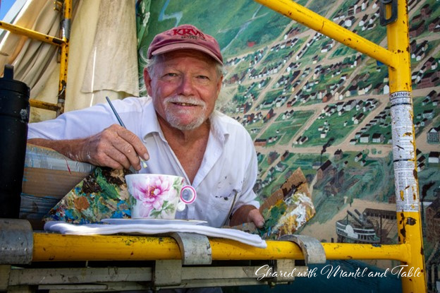 Robert Dafford, renowned artist, on scaffolding, painting murals. Teacup and saucer in the foreground.