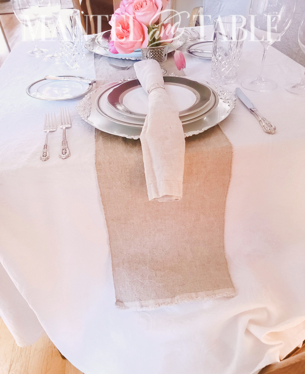 Silver and charcoal place setting on a table with a long white tablecloth and tan linen runner