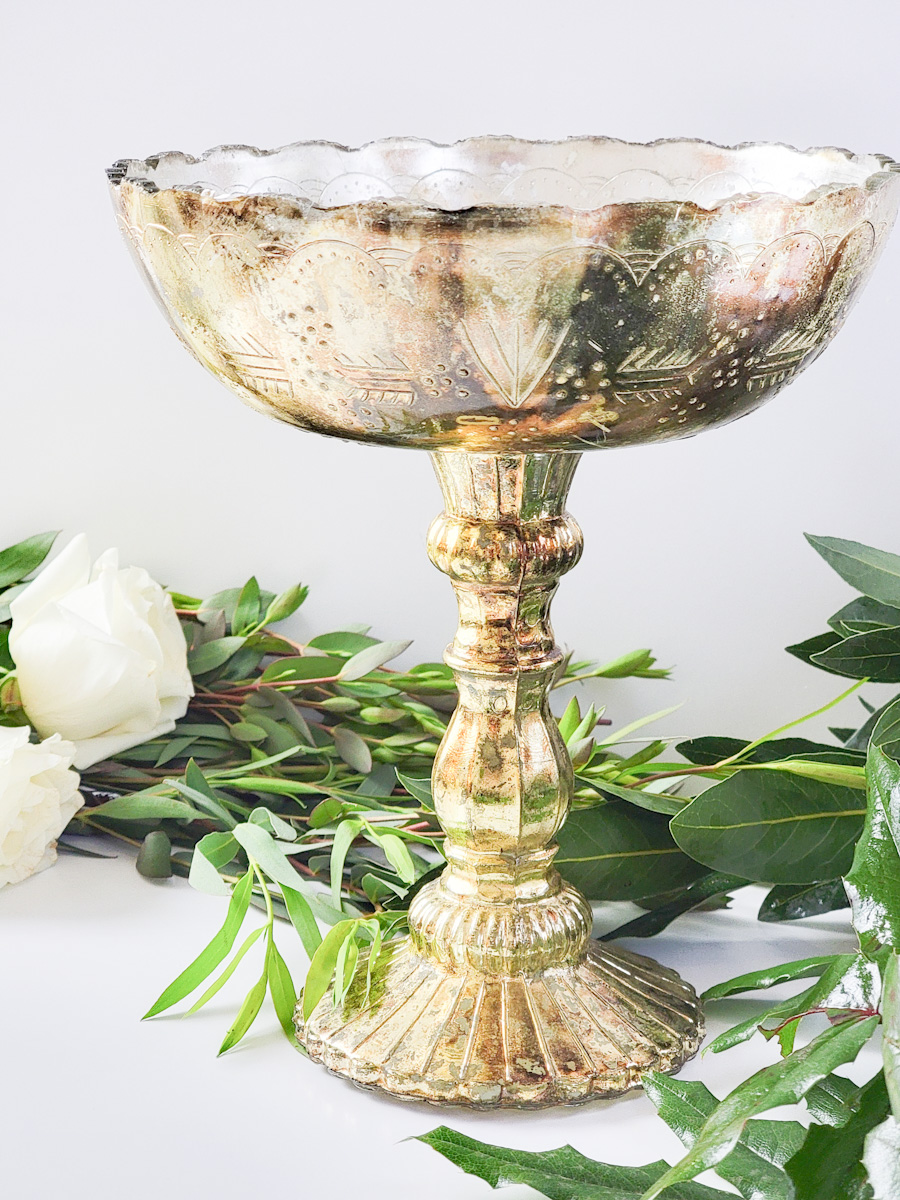 Closeup of a gold mercury glass pedestal vase with greenery around it