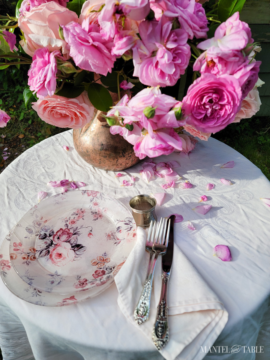 copper pitcher of pink roses on a table with pink tablecloth and fallen petals